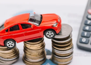 A customer signing a vehicle finance agreement at a dealership desk with a new car in the showroom behind