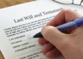 Hand signing a printed last will and testament at a desk while two witnesses look on