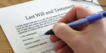 Hand signing a printed last will and testament at a desk while two witnesses look on