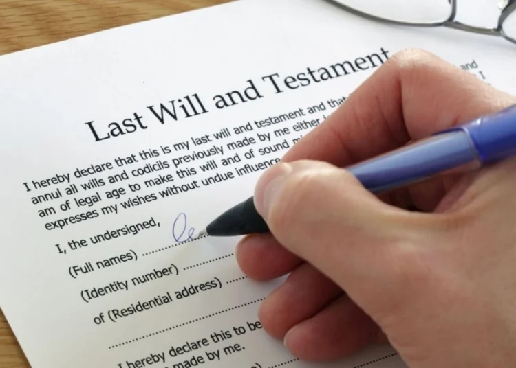 Hand signing a printed last will and testament at a desk while two witnesses look on